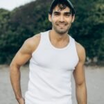 Casually dressed man smiling outdoors on a sandy beach with greenery.